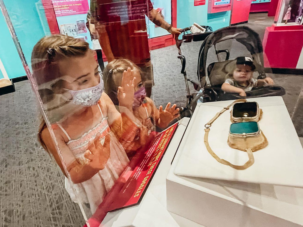 Family wearing face masks looking at Amelia Earhart's aviator goggles in the Barbie exhibit.